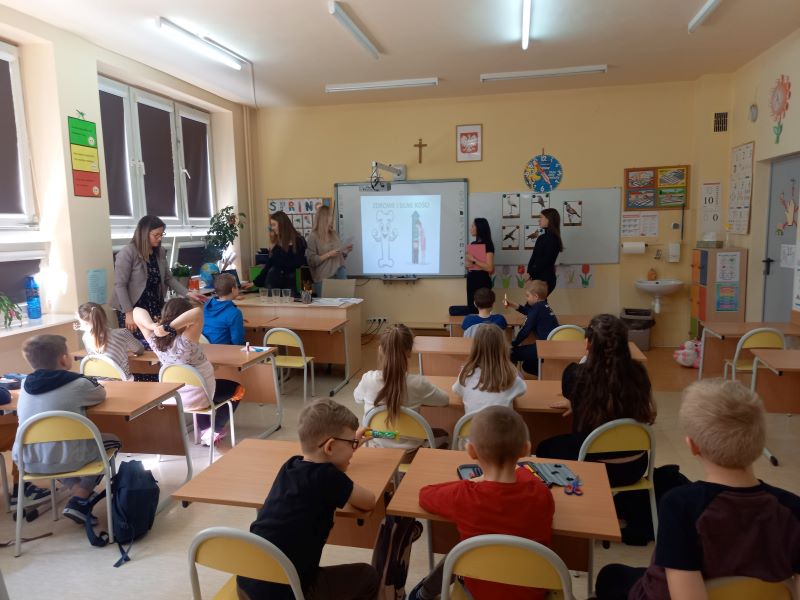 w sali siedzą dzieci w ławkach. 5 Pań daje instrukcje. Widać projektor, krzyż, godło Polski. Na ścianach obrazki
