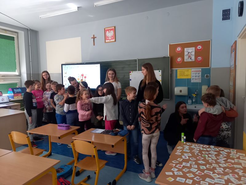dzieci stoją w sali. Za nimi są 3 kobiety. Widać godło Polski i krzyż. Na ekranie jest jakiś obraz