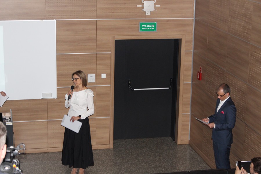 dr J. Klusek przemawia z mikrofonem. Prof. M. Sadowski czyta dokument. Obydwoje stoją w auli. Widać drzwi