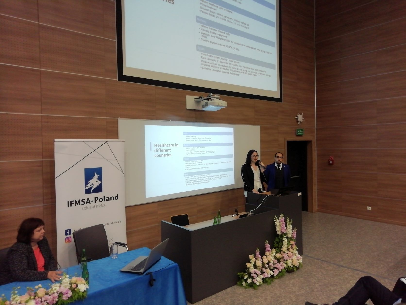 przy stole na auli siedzi dr Brzozowska. Studenci Daniel Minguez Gala i Anesa Ahmatovic prezentują.