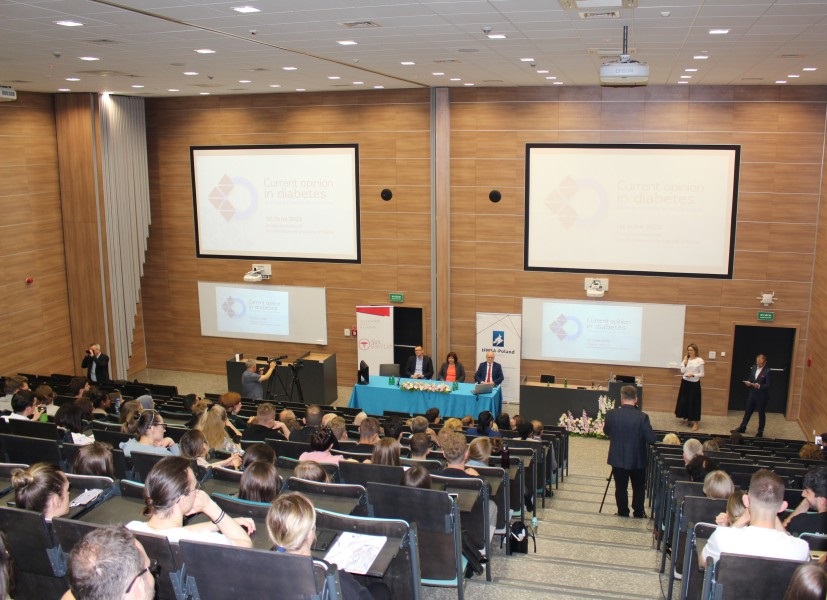 na auli widać uczestników konferencji. Widać obsługę techniczną. Widać 4 ekrany. Widać stół za, którym siedzą prelegenci konferencji